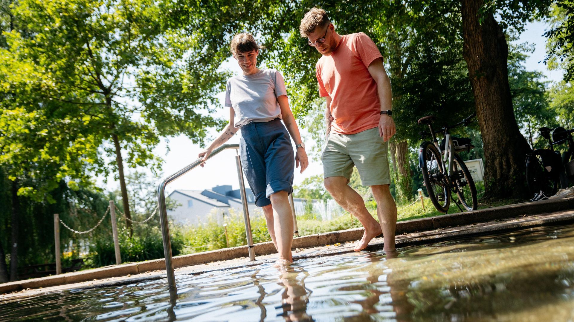 Ein Paar kühlt barfuß ihre Füße in einem Wasserbecken unter schattigen Bäumen.