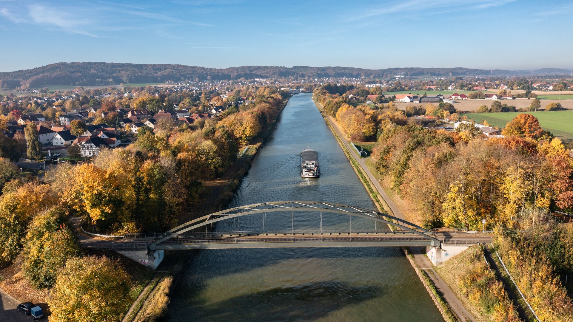 Brücke über einen breiten Kanal mit einem Schiff, umgeben von herbstlichen Bäumen.
