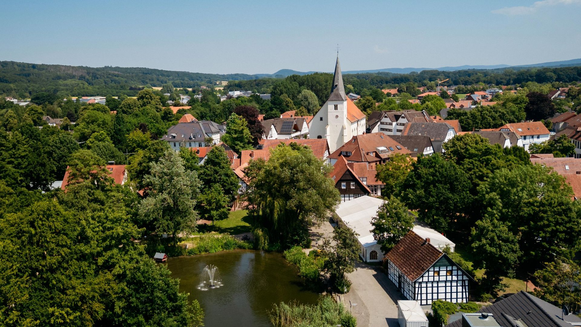 Kirche mit spitzem Turm in grüner Landschaft, umgeben von Gebäuden und kleinem Teich.