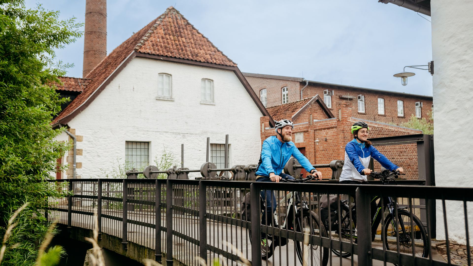 Zwei Radfahrer überqueren eine Brücke vor einem historischen Gebäude mit Schornstein.