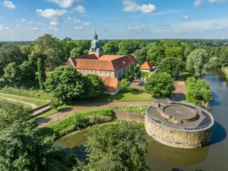 Historisches Schloss inmitten üppiger grüner Landschaft mit Wassergraben und Rundturm im Vordergrund.