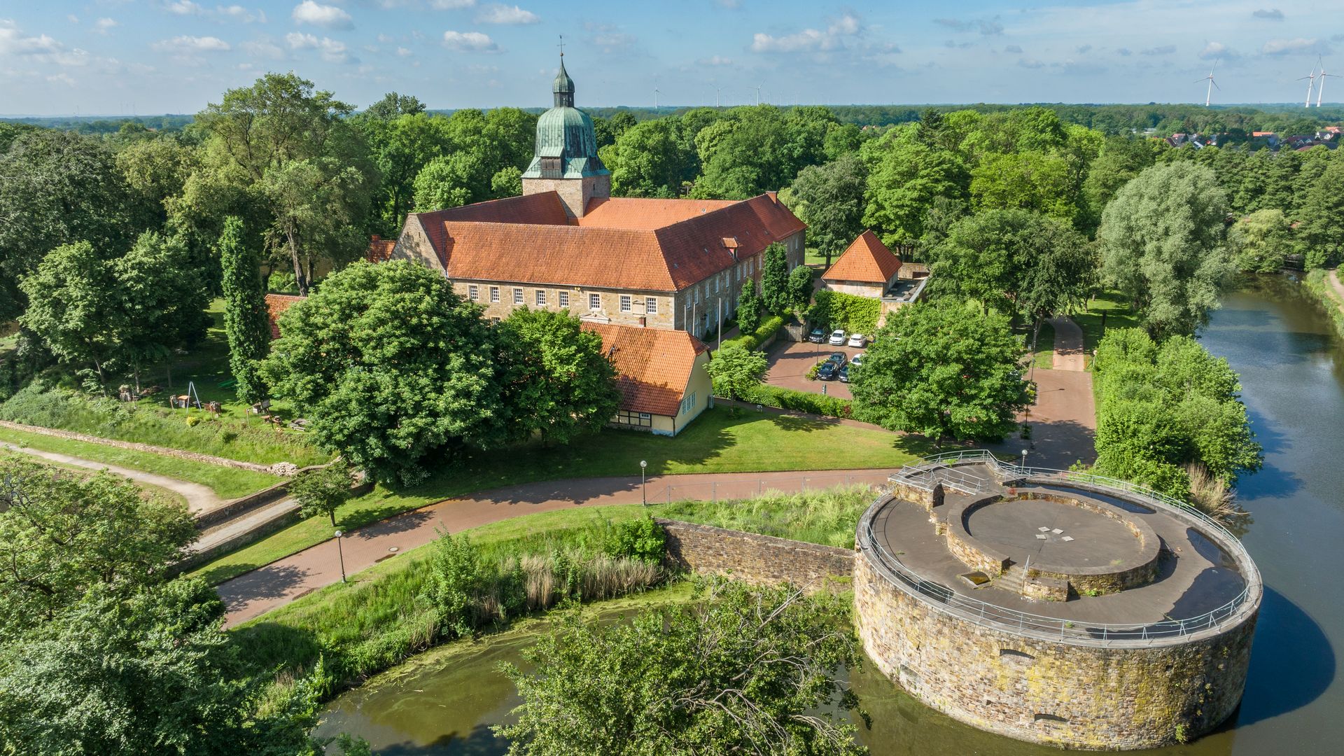 Historisches Schloss inmitten üppiger grüner Landschaft mit Wassergraben und Rundturm im Vordergrund.