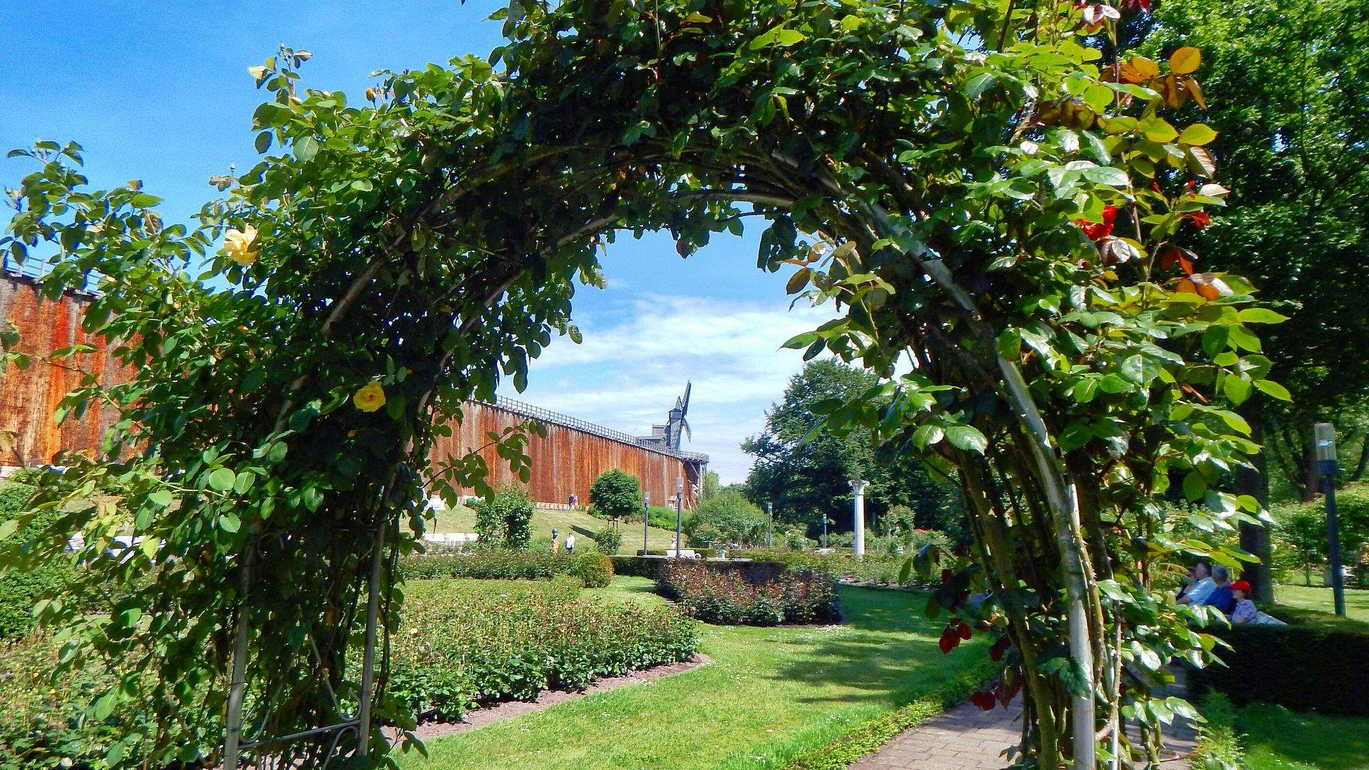 Rosenbogen in einem gepflegten Garten mit sattem Grün und blauem Himmel im Hintergrund.