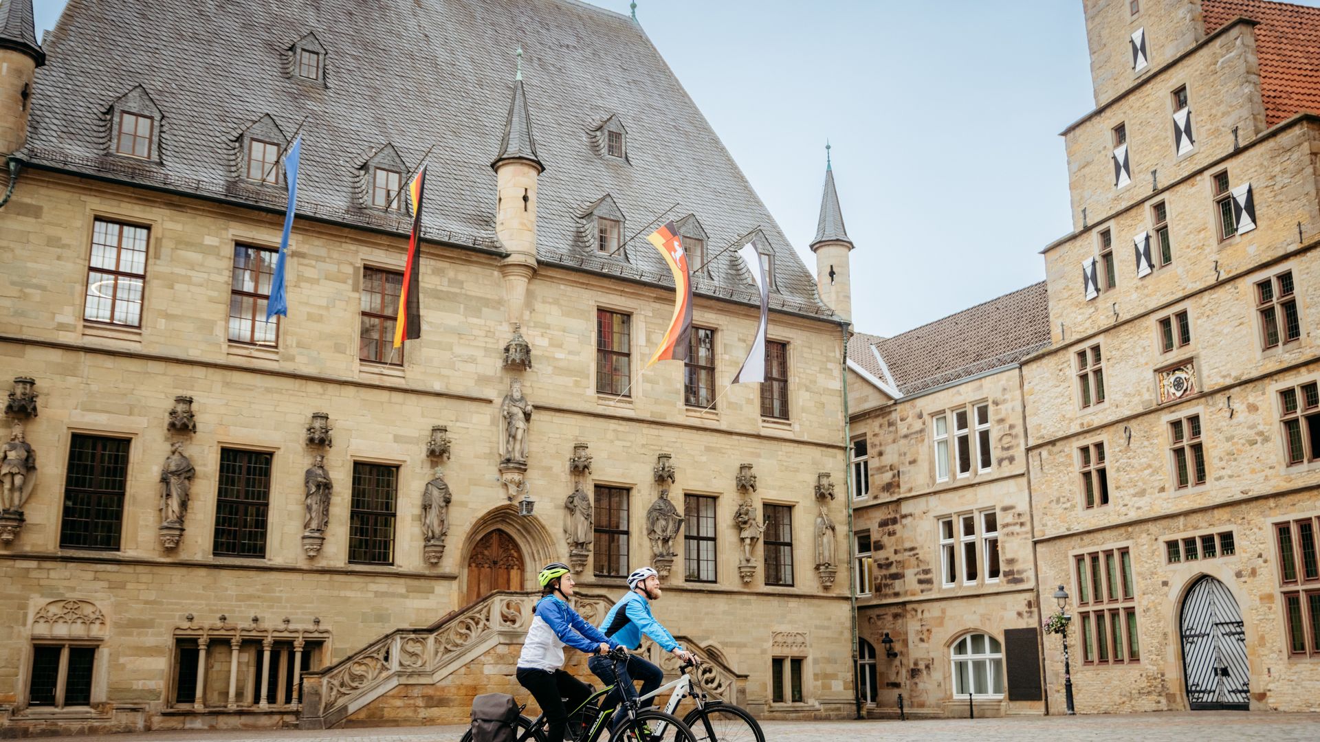Zwei Radfahrer vor einem historischen Sandsteingebäude mit Skulpturen.