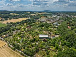 Luftaufnahme einer weitläufigen Zooanlage, umgeben von Grünflächen und Wäldern, unter einem blauen Himmel.