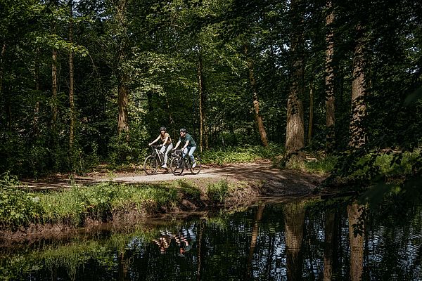 Zwei Personen fahren mit ihren Fahrrädern an einem Fluss entlang 