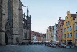 Abendliche Marktplatzszene mit beleuchteten Gebäuden und Menschen, die an Tischen im Freien sitzen.