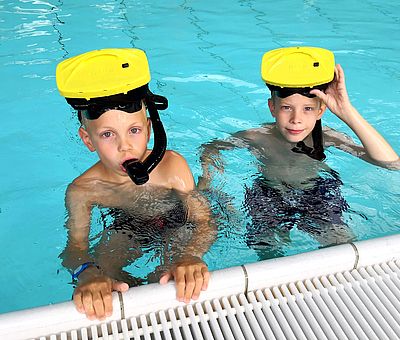 Auf diesem Bild geht es um das VR-Schnorcheln im Nettebad Osnabrück. Zwei kleine Jungen stehen mit der VR-Brille auf dem Kopf und dem Schnorchel im Wasser am Beckenrand. Der linke Junge hält sich mit beiden Händen am Wasserrand fest und hat seinen Schnorchel im Mund. Der rechte Junge hält seine VR-Brille mit der einer Hand an seinem Kopf fest. 