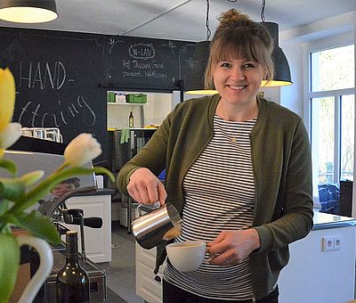 Im Ausflugslokal Hof Thiesing in Osnabrück gibt es vieles zu sehen und leckeren Kaffee. Auf diesem Foto steht eine Frau, die gerade dabei ist, Kaffee in eine weiße Tasse zu gießen. Neben der Frau steht eine Blumenvase mit bunten Tulpen. Die Wand hinter der Frau ist schwarz gestrichen und es ist mit einer weißen Schrift etwas drauf geschrieben, was man leider nicht lesen kann. Links hinter den Tulpen steht eine große silberne Kaffeemaschine. 