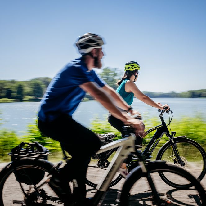 Zwei Radfahrer genießen eine Fahrt entlang eines ruhigen Sees an einem sonnigen Tag.