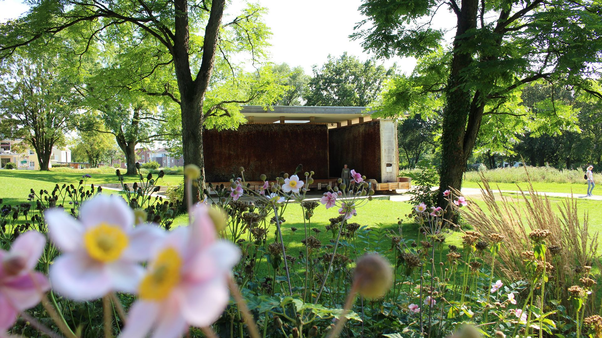 Rostfarbene Stahlwand in einem sonnigen Park, umgeben von blühenden Blumen und Bäumen.