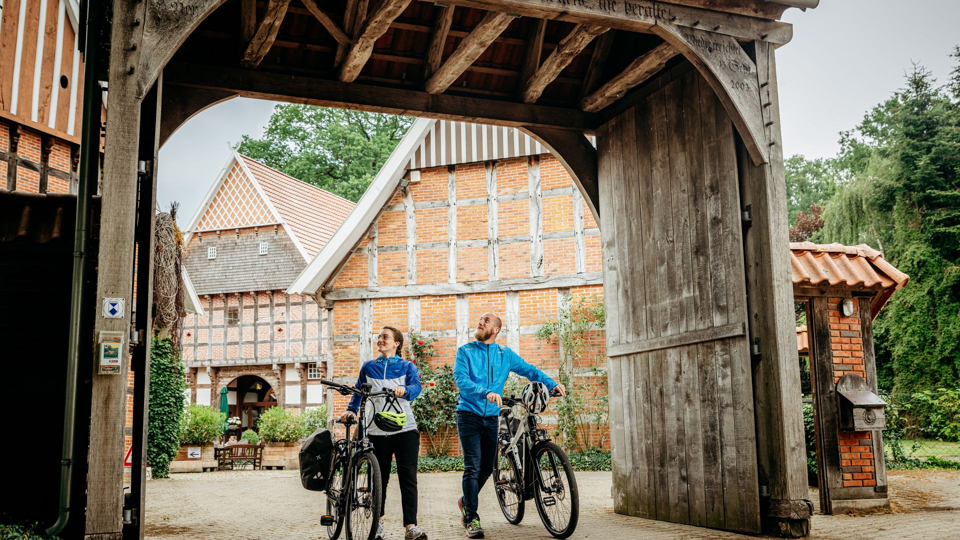 Zwei Radfahrer stehen unter einem hölzernen Torbogen, umgeben von Fachwerkhäusern.
