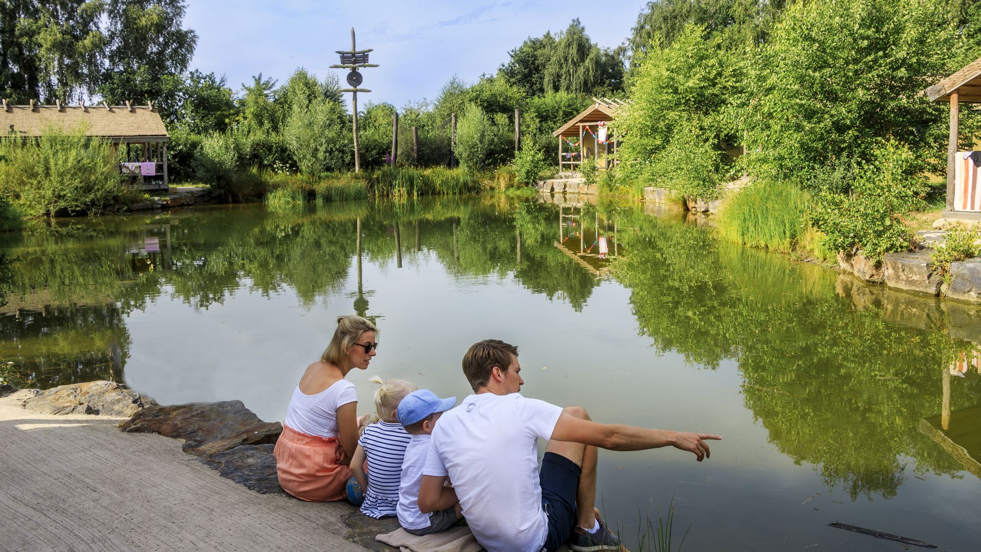 Familie am Teich auf Steinmauer sitzend, umgeben von Bäumen, beobachtet das Wasser.