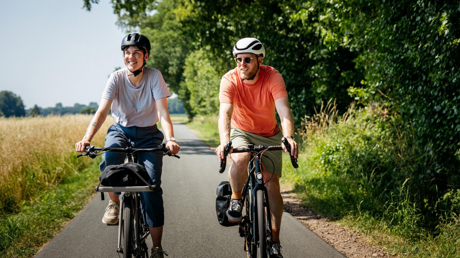 Zwei Radfahrer auf einem asphaltierten Weg im Grünen, lächelnd unterwegs.