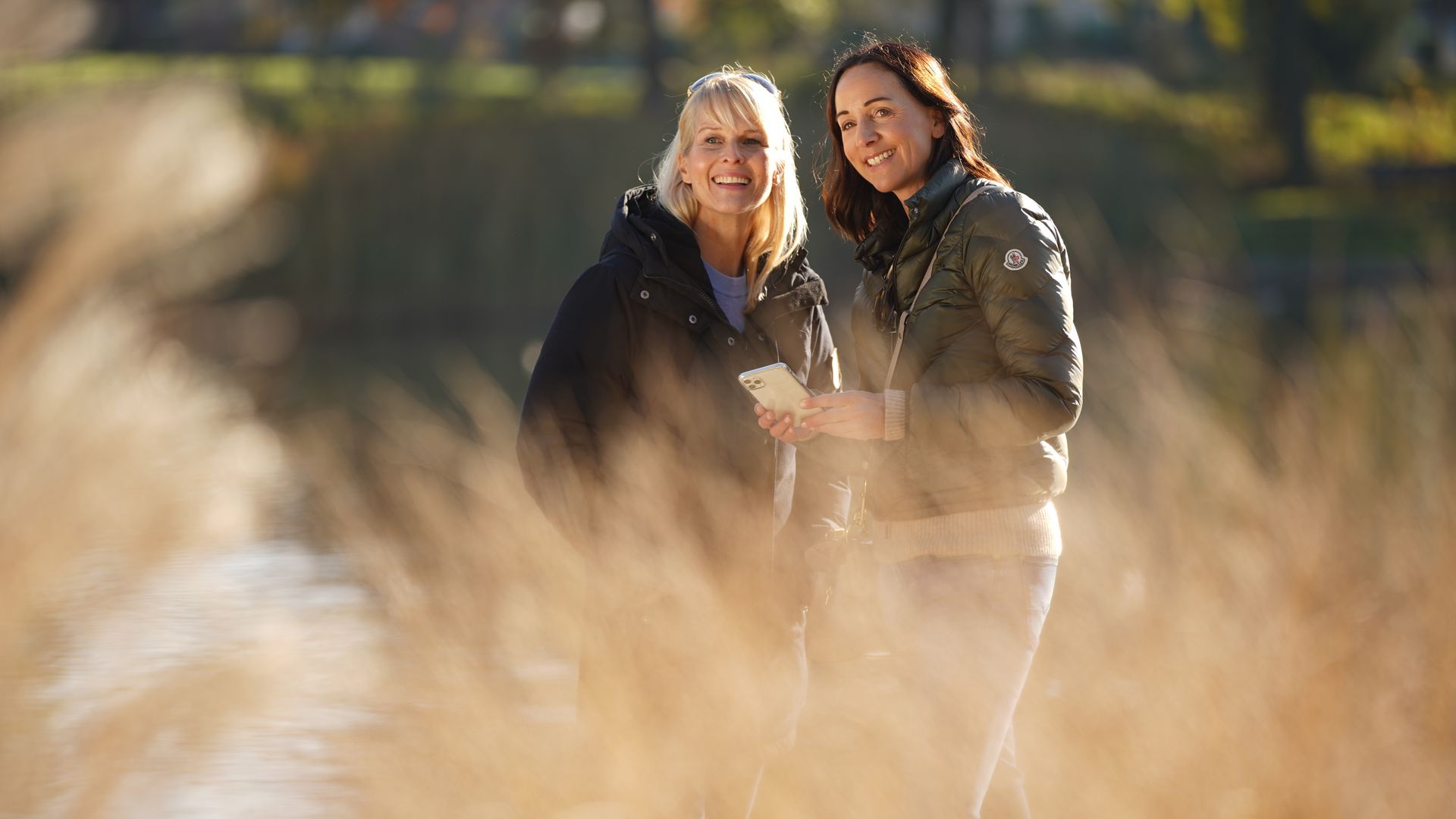 Zwei Frauen stehen lächelnd in einem sonnigen Park vor verschwommenem Gras.