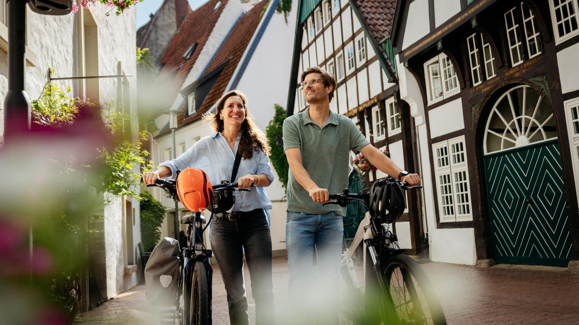 Zwei Personen laufen mit Fahrrädern durch eine gepflasterte Straße mit Fachwerkhäusern.