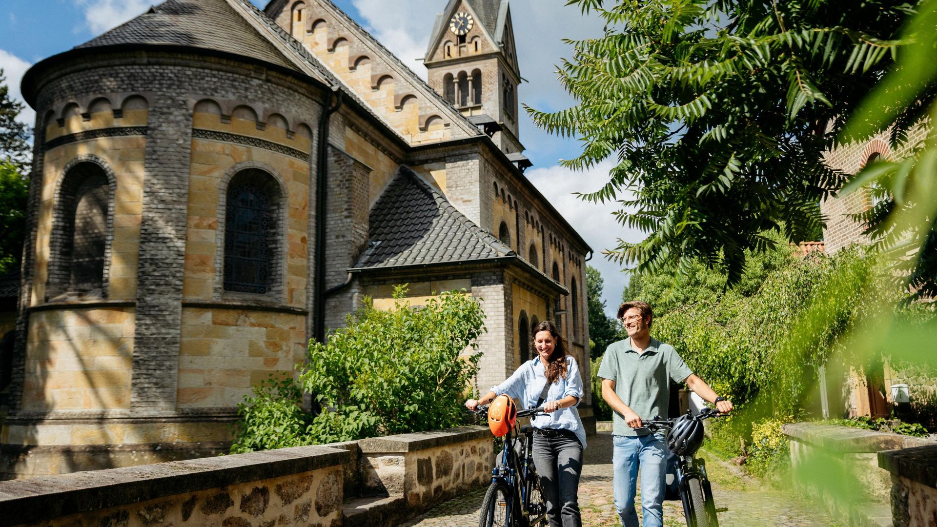 Zwei Radfahrer vor einer alten Backsteinkirche bei sonnigem Wetter in einer grünen Umgebung.