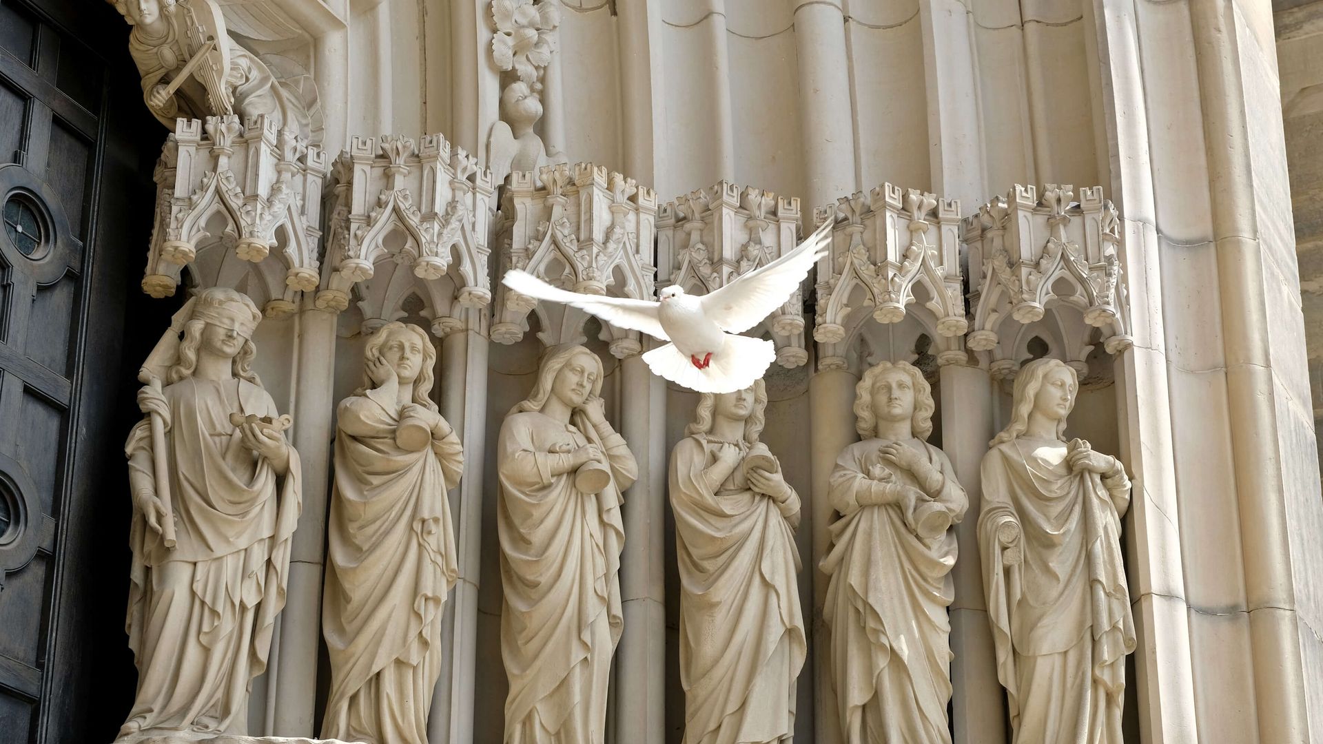 Eine weiße Taube fliegt vor einer gotischen Steinreliefdarstellung mit Heiligen.