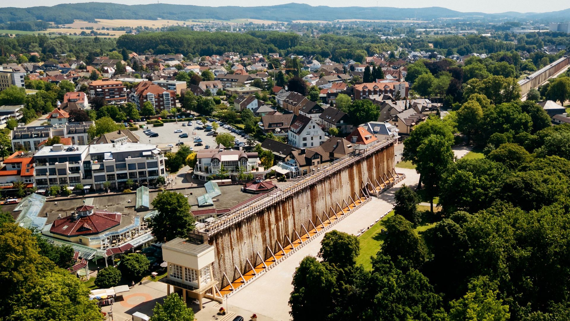 Gradierwerk in einer grünen Parklandschaft, umgeben von Stadthäusern und Hügeln im Hintergrund.