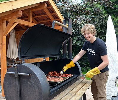 Im Ausflugslokal Hof Thiesing in Osnabrück wird auch fleißig für die Gäste gegrillt. Auf diesem Foto steht ein junger Mann mit blonden Locken vor dem Grill und lächelt in die Kamera. Er trägt gelbe große Handschuhe und dreht gerade mit einer Zange das Fleisch auf dem Grill um. Der Grill ist tonnenförmig und hat eine Klappe, die man zum Grillen öffnet. Ringsrum stehen Bäume und links auf dem Bild hinter dem Grill sieht man einen Holzschuppen. 