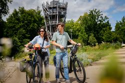 Ein Paar fährt lächelnd auf E-Bikes an einem hohen Aussichtsturm vorbei.