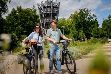 Ein Paar fährt lächelnd auf E-Bikes an einem hohen Aussichtsturm vorbei.
