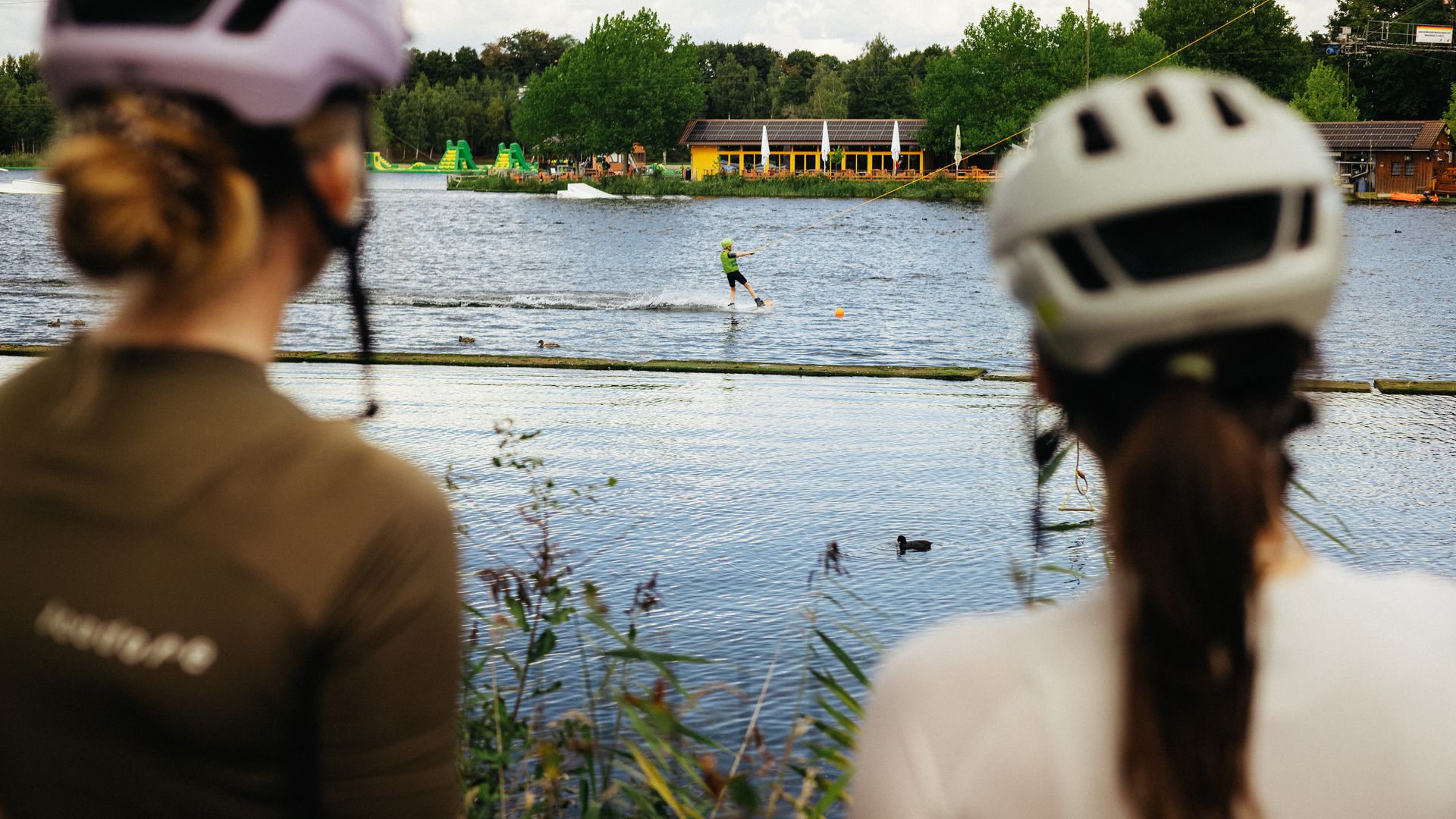 Zwei Rennradfahrende mit Helm blicken vom Ufer auf den Alfsee, während Wassersport auf dem See stattfindet.