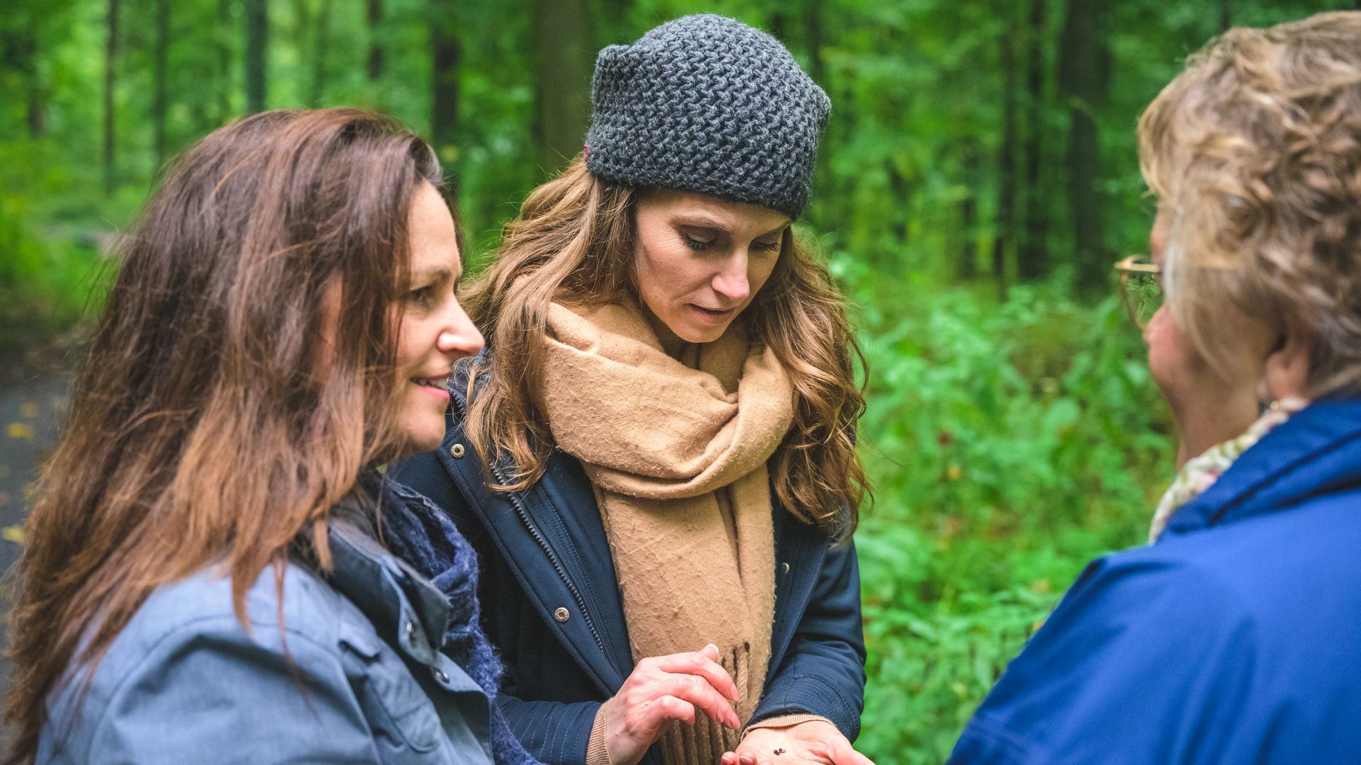 Drei Frauen unterhalten sich in einem Wald, während eine von ihnen etwas in der Hand hält.
