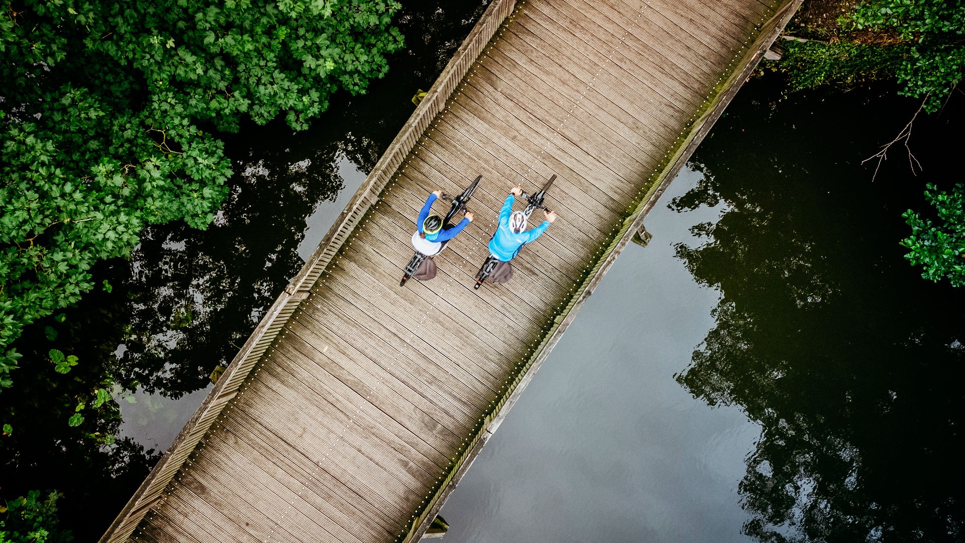 Zwei Radfahrer auf einer Holzbrücke über einem ruhigen Fluss, umgeben von grünen Bäumen.