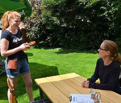 Das Foto wurde aufgenommen auf Hof Thiesing, einem Ausflugslokal in Osnabrück. Eine junge rothaarige Frau, die ein schwarzes T-Shirt mit einem weißen Logo von Hof Thiesing trägt, bedient eine andere Frau, die im Biergarten an einen Holztisch sitzt. Auf dem Tisch liegt eine Speisekarte & eine Tischnummer mit der Nummer vier.