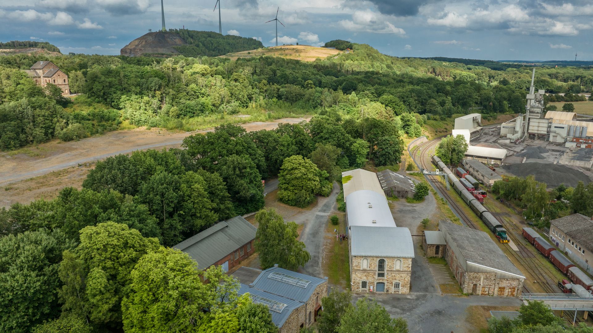 Ehemaliges Industriegelände mit Zügen und Windturbinen in grüner Hügellandschaft.