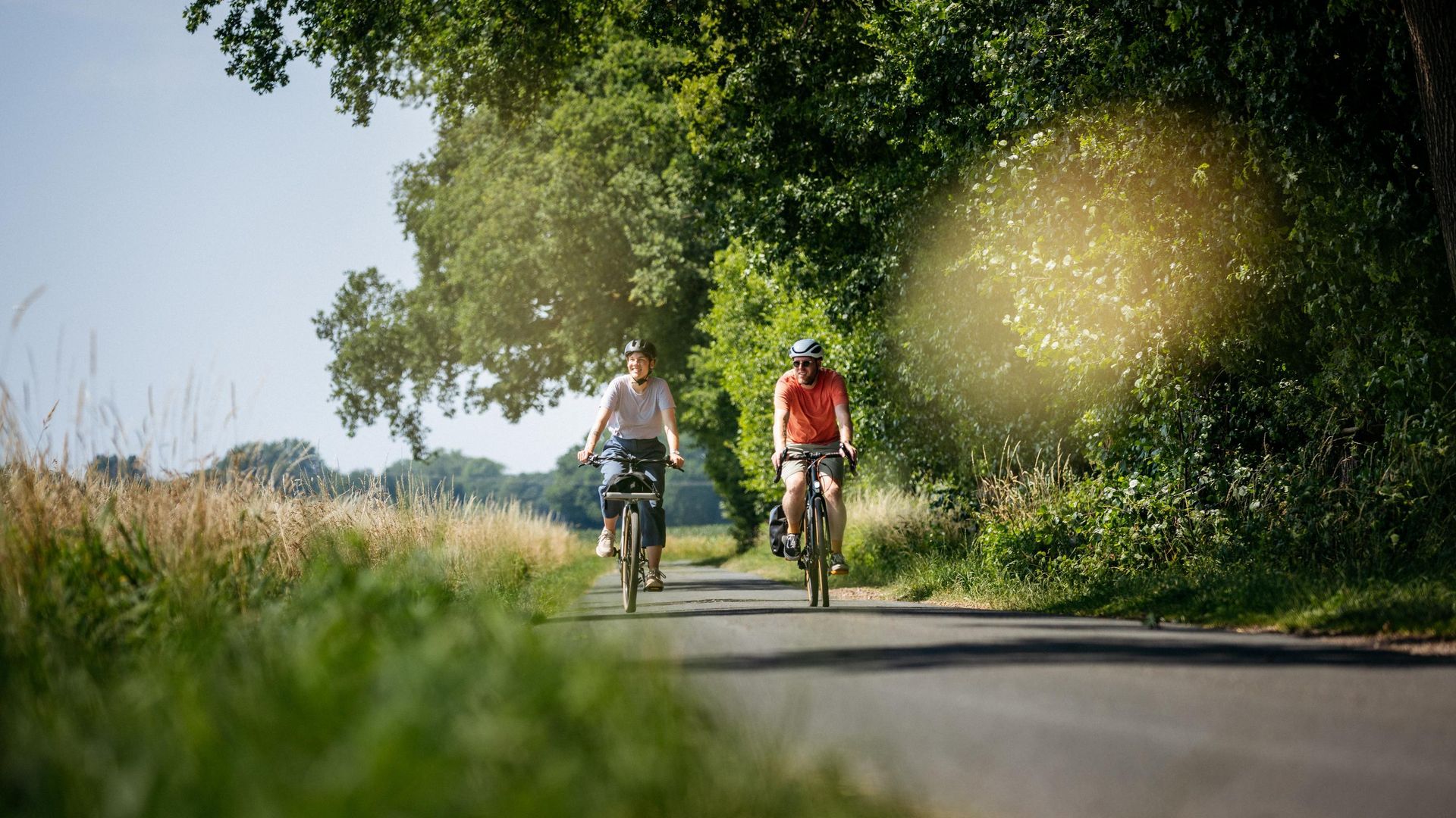 Zwei Radfahrer fahren auf einer Landstraße, umgeben von grüner Natur.