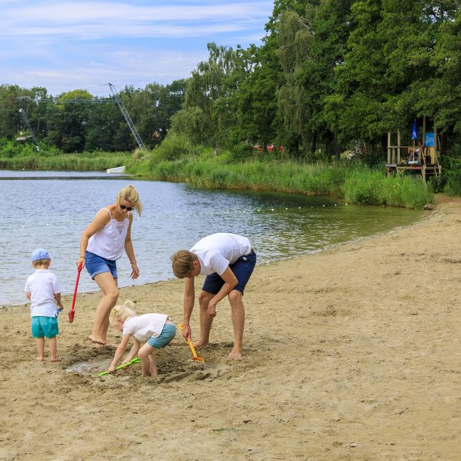 Familie spielt gemeinsam am sandigen Ufer eines Sees, umgeben von grünen Bäumen.