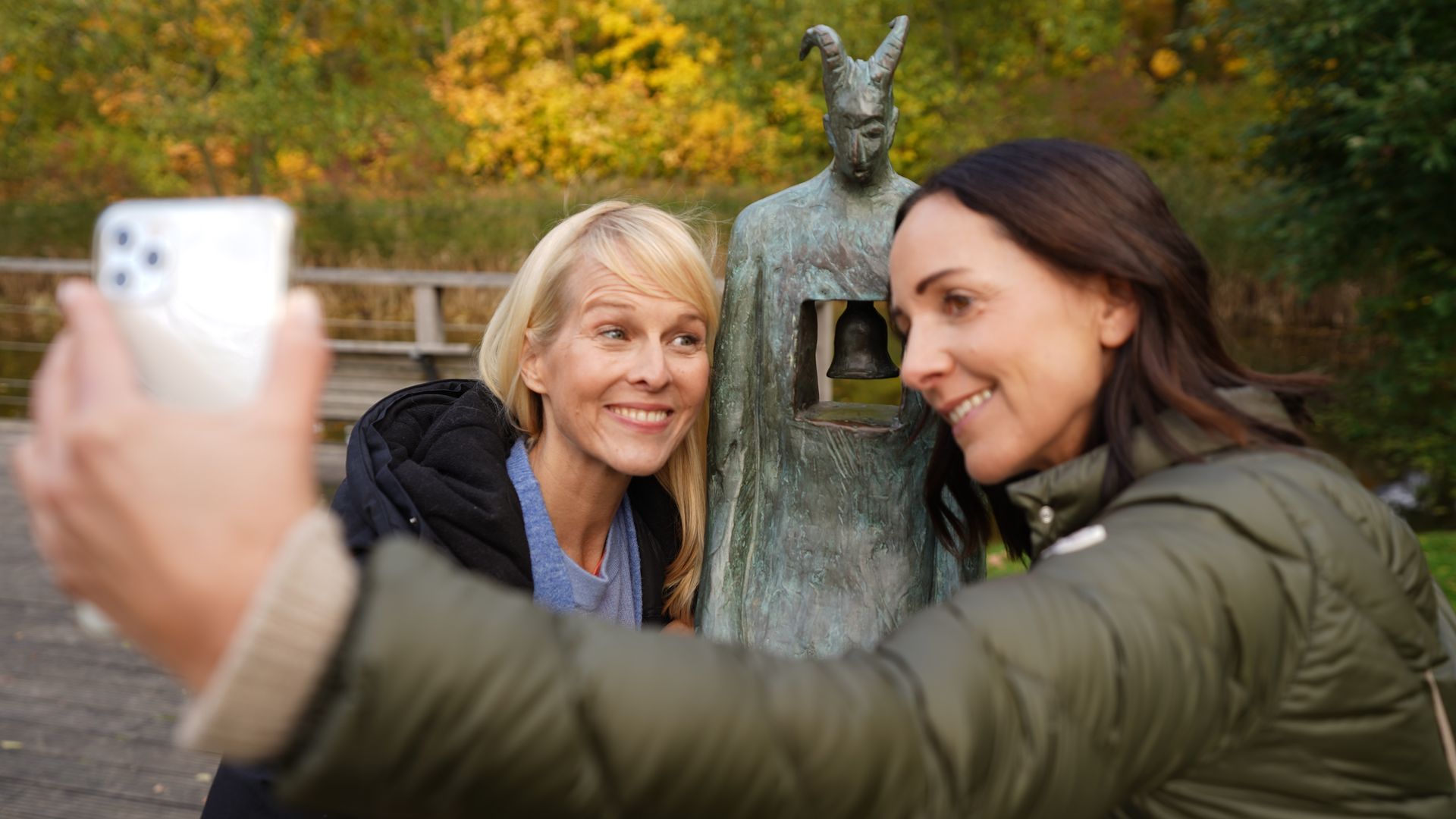 Zwei Frauen machen ein Selfie mit einer Bronzeskulptur in einem herbstlichen Park.