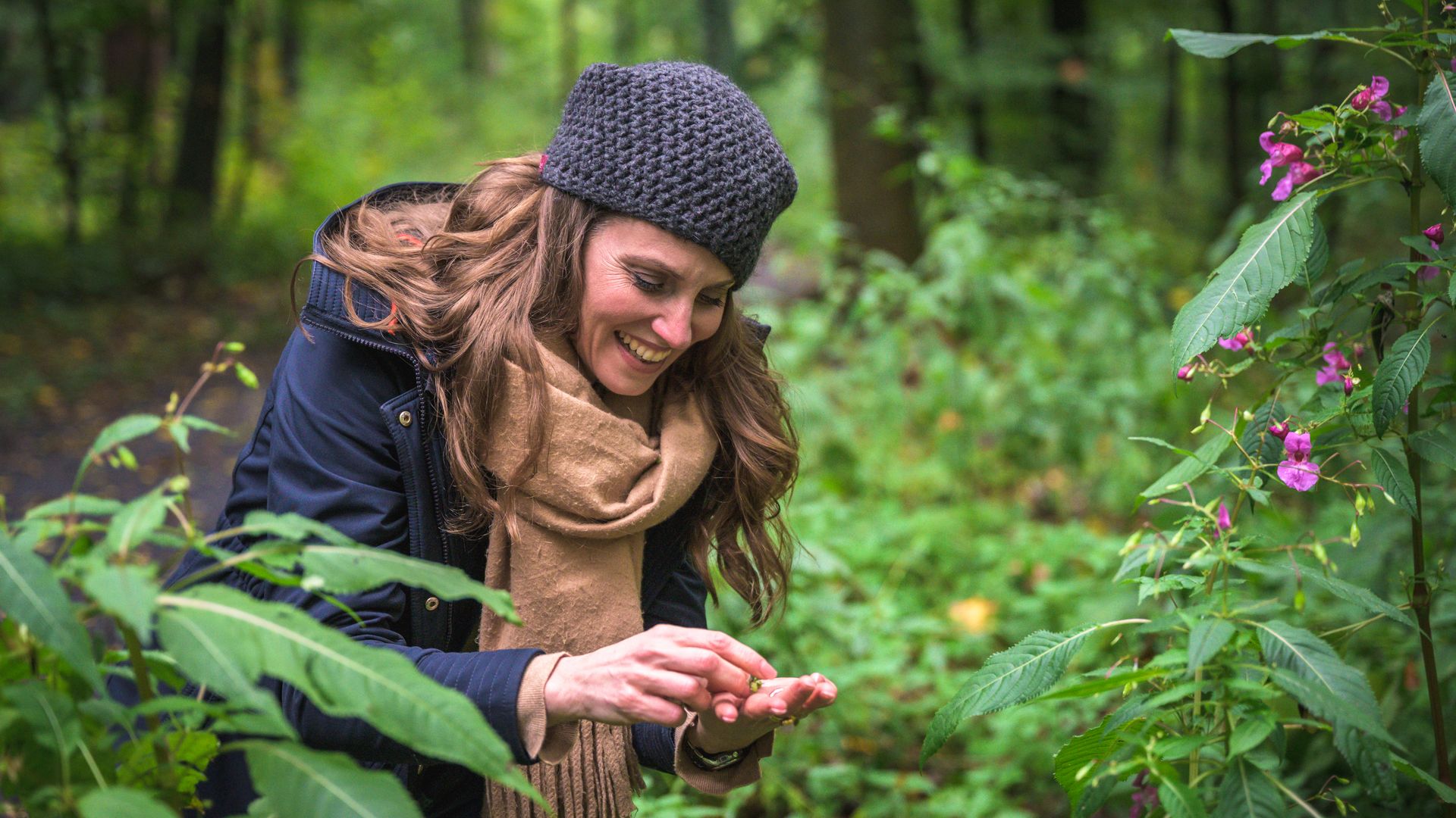 Frau lächelnd im Wald, untersucht eine Pflanze mit pinken Blüten neben einem bewachsenen Pfad.