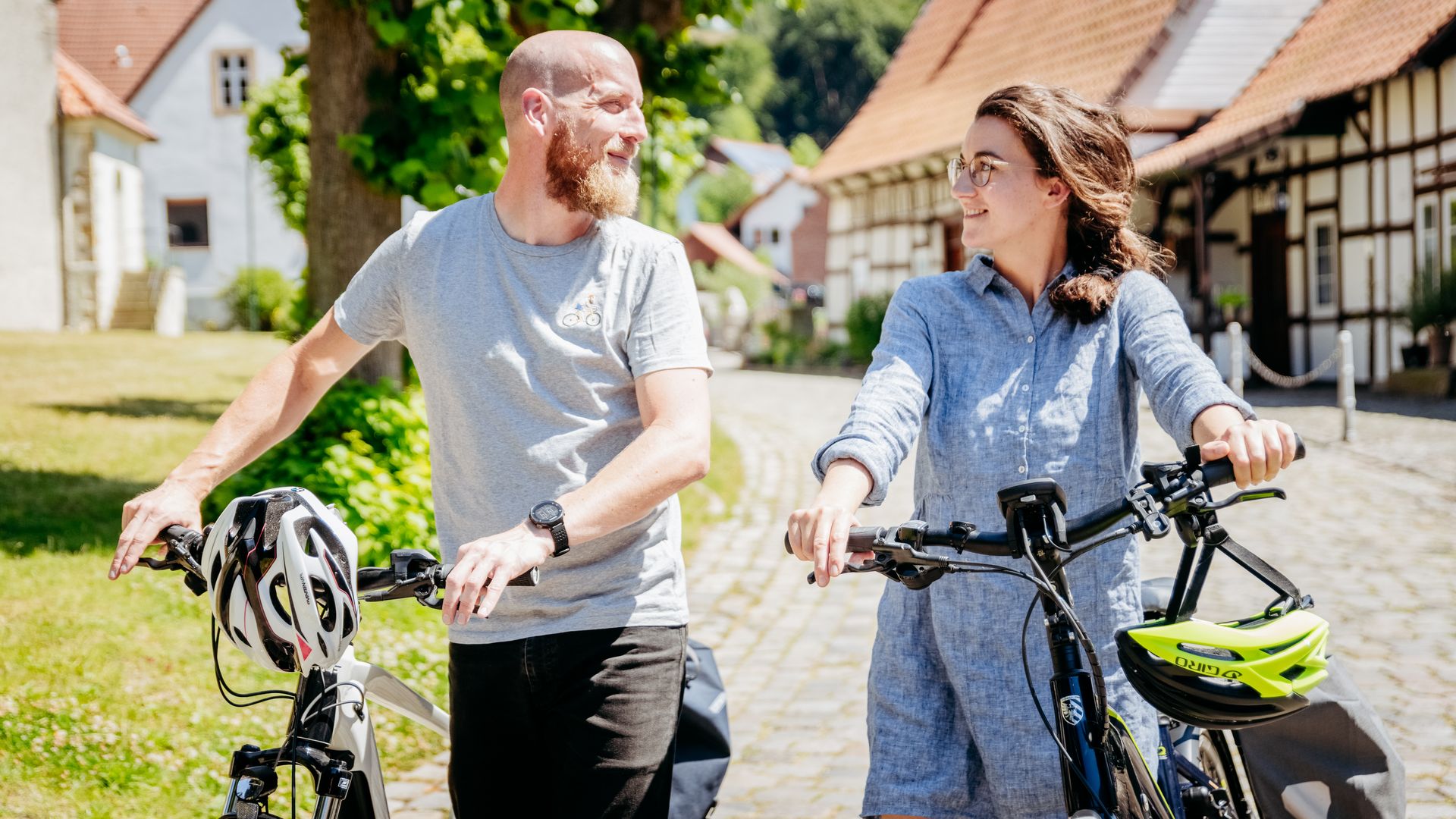 Zwei Personen schieben Fahrräder durch ein sonniges Dorf mit Fachwerkhäusern.