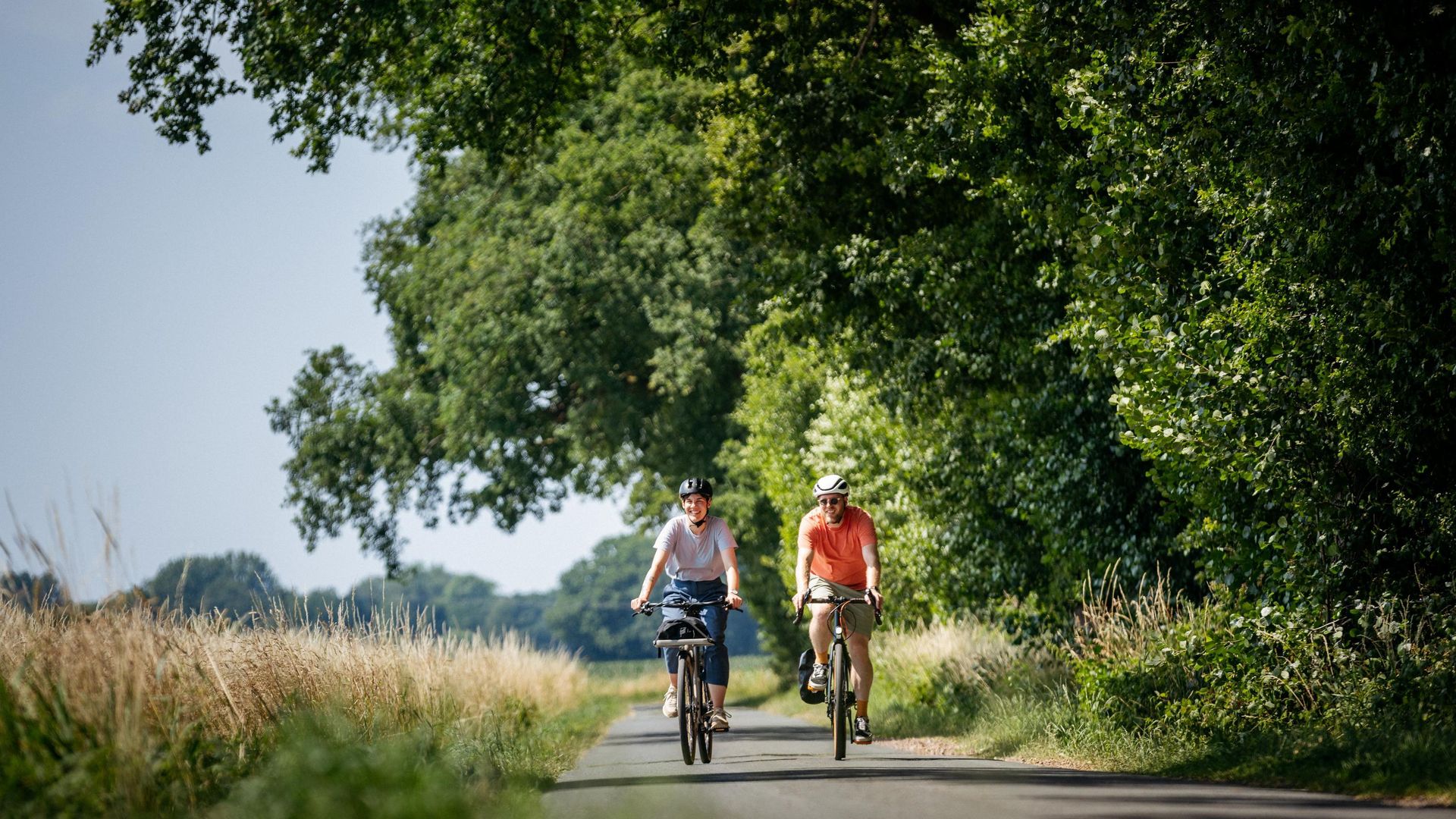 Zwei Personen radeln entspannt auf einem von Bäumen gesäumten Landweg an einem sonnigen Tag.