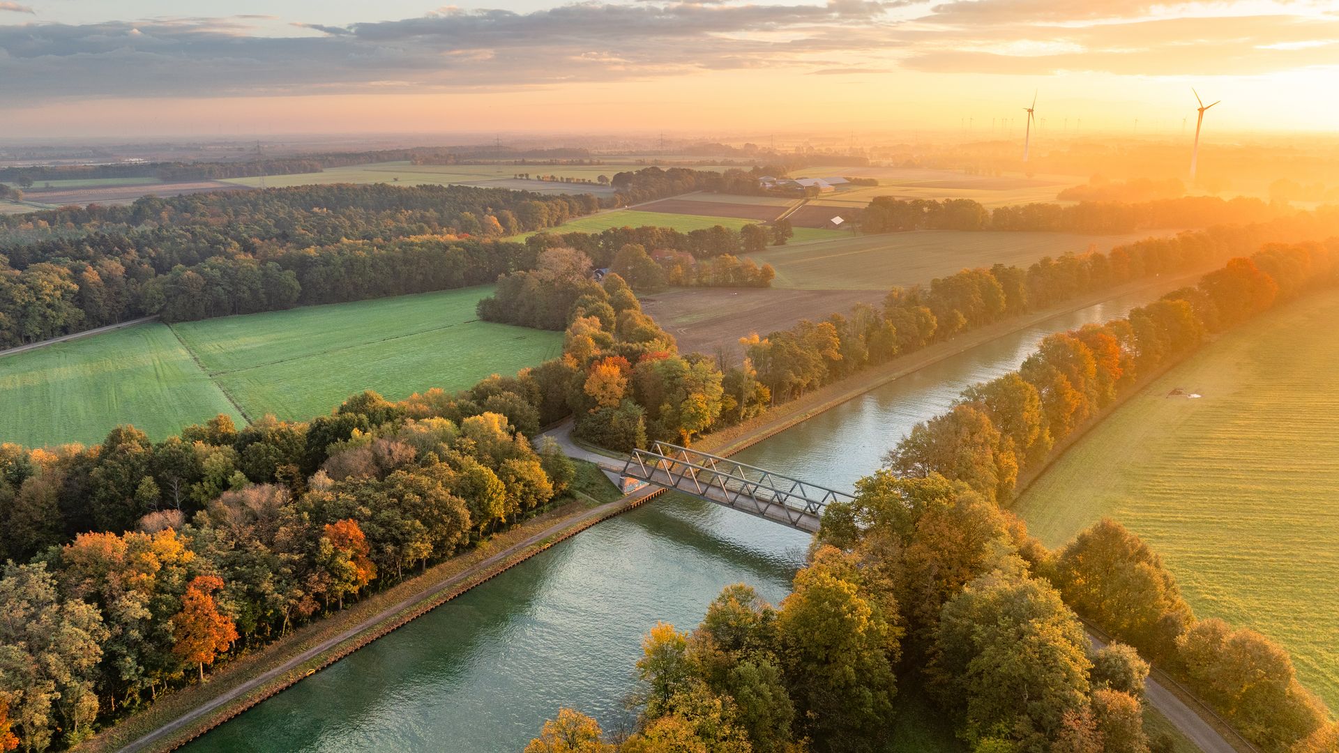 Luftaufnahme eines Kanals mit Brücke, umgeben von herbstlich gefärbten Bäumen bei Sonnenuntergang.