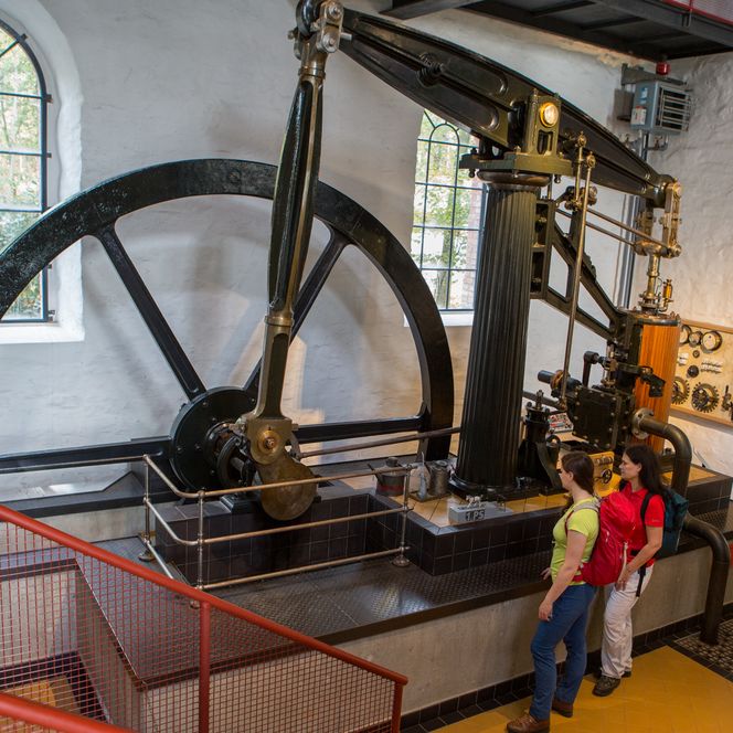 Zwei Personen betrachten eine historische Dampfmaschine mit Schwungrad in einem Industriemuseum.