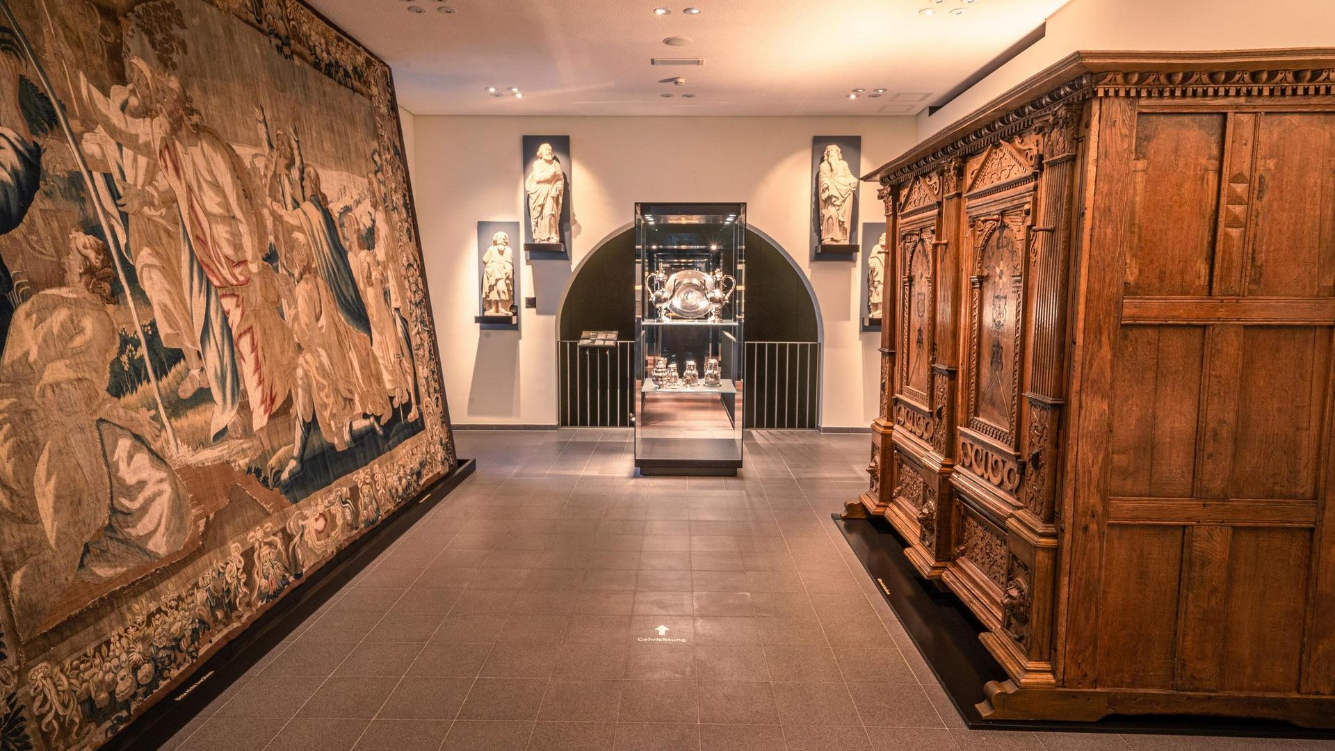 Ausstellungsraum im Diözesanmuseum Osnabrück mit Skulpturen, wertvollem Wandteppich und Holzschrank.