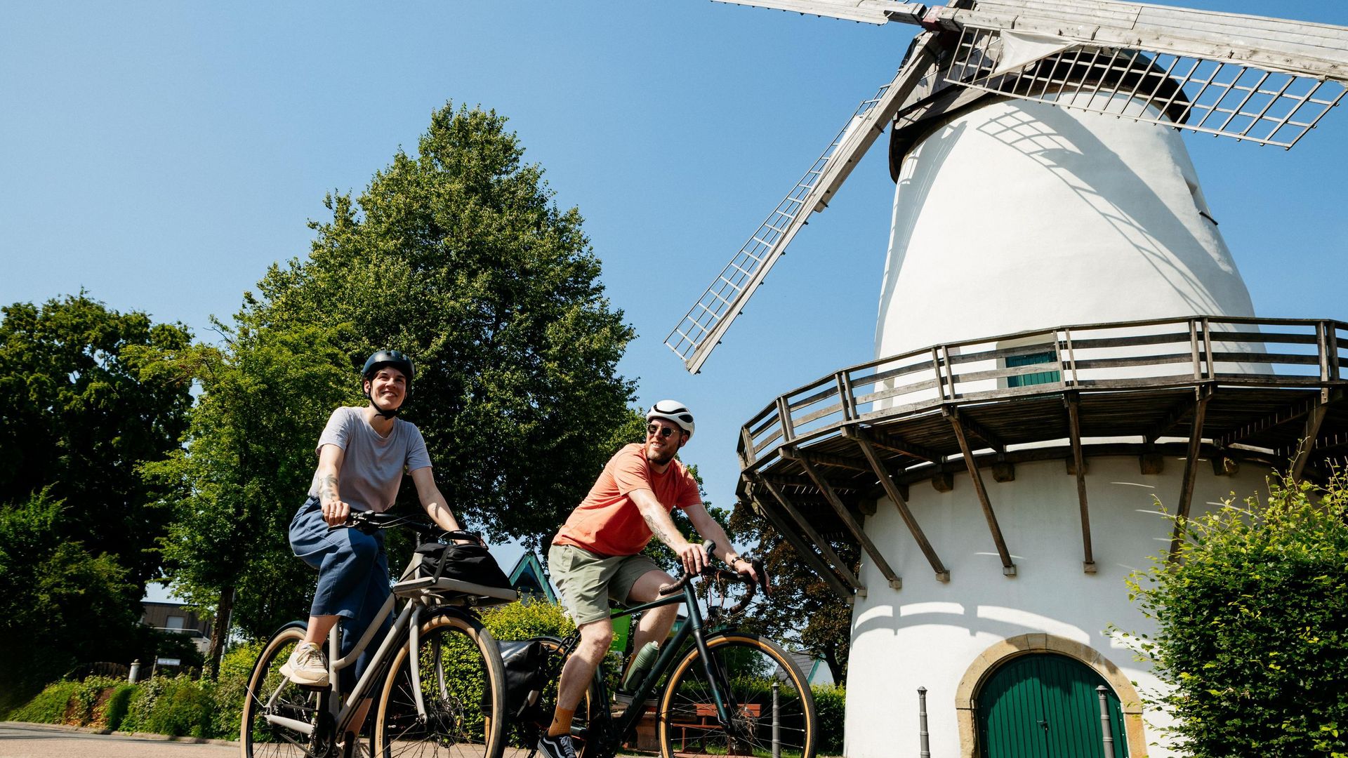 Zwei Radfahrer vor einer historischen Windmühle bei sonnigem Wetter.