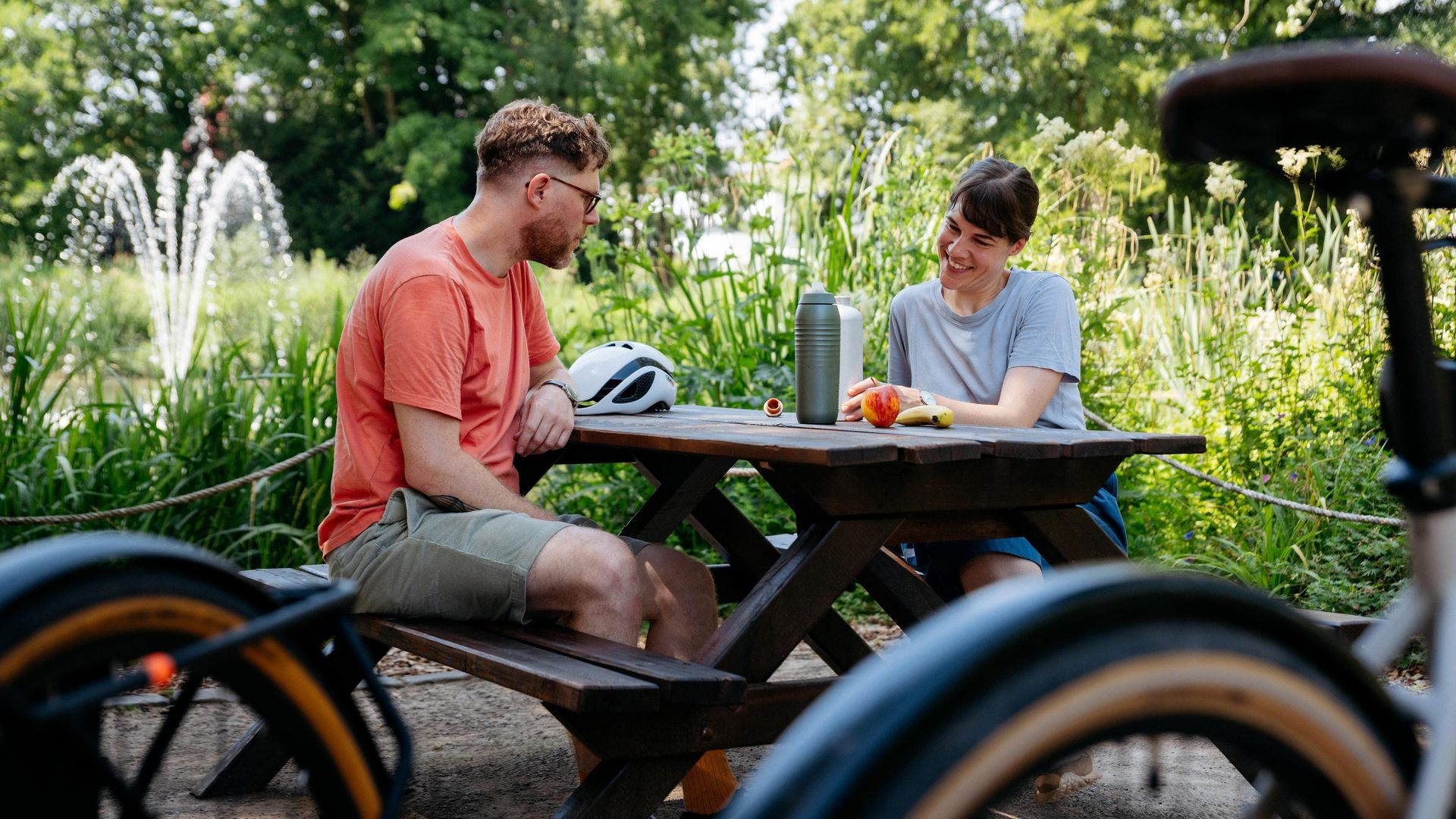 Zwei Personen sitzen an einem Picknicktisch im Grünen, umgeben von Fahrrädern und Pflanzen.