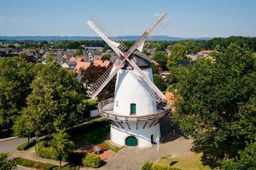 Windmühle mit rotierenden Flügeln inmitten grüner Landschaft und kleiner Häuser.