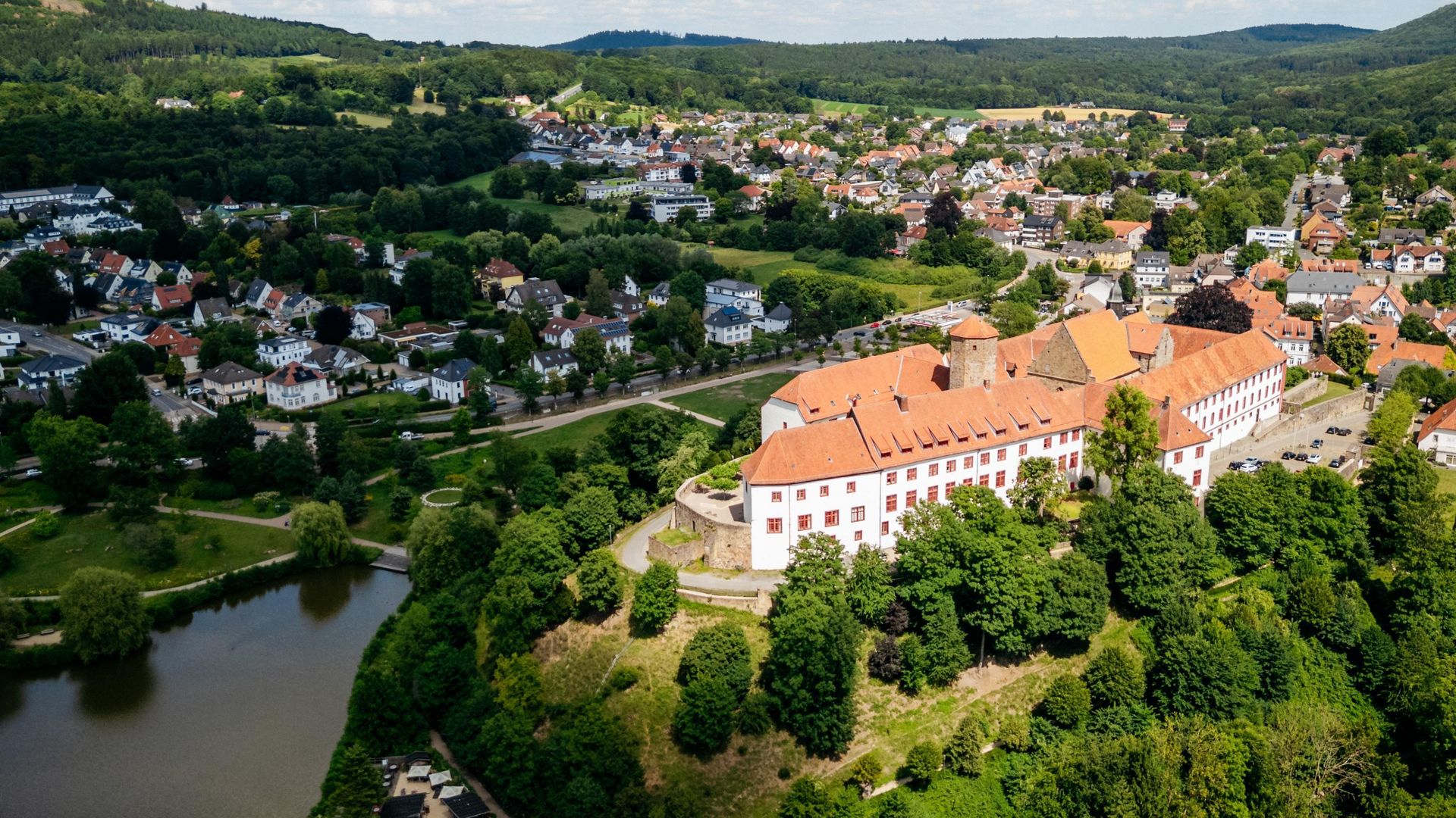 Luftaufnahme einer großen, historischen Burg umgeben von grünen Hügeln und einem See.