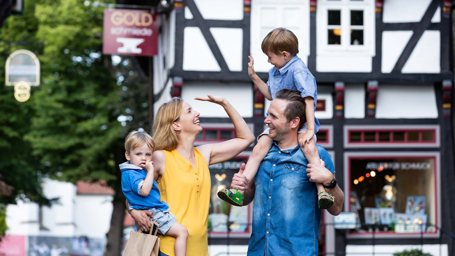 Eine fröhliche Familie steht vor einem Fachwerkhaus im Osnabrücker Land. Die Mutter und der Vater lachen, während der Vater den älteren Sohn auf den Schultern trägt und die jüngere Tochter von der Mutter gehalten wird. Das historische Gebäude im Hintergrund verleiht der Szene einen charmanten, regionalen Charakter.