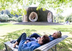 Zwei Frauen entspannen auf einer Bank vor einem großen, skulpturalen Brunnen im Park.