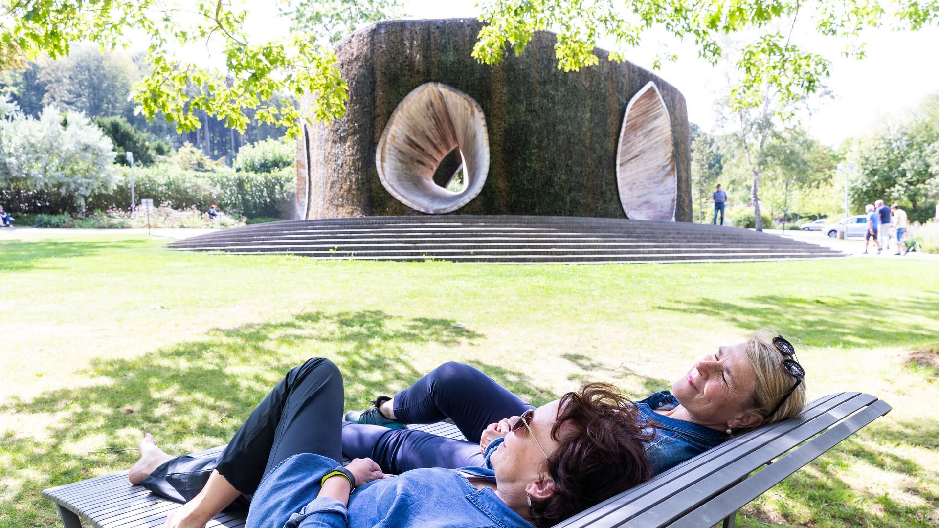 Zwei Frauen entspannen auf einer Bank vor einem großen, skulpturalen Brunnen im Park.