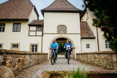 Zwei Radfahrer passieren ein historisches Torhaus auf Kopfsteinpflaster.