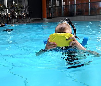 Auf diesem Foto bekommt man einen Einblick, wie das VR-Schnorcheln im Nettebad Osnabrück aussehen kann. Im Schwimmbecken ist ein kleiner blonder Junge zu sehen, der gerade dabei ist zu schnorcheln. Mit seiner gelben VR-Brille, die er mit beiden Händen auf seinen Kopf trägt, kann er sich in eine andere Welt versetzen.