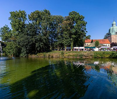 Dieses Foto ist beim Sommerfest der Kulturen in Fürstenau im Osnabrücker Land entstanden. Im Bildvordergrund ist Wasser mit einer kleinen Wasserfontäne zu sehen, am Ufer dahinter ist die Schlossinsel von Fürstenau zu erkennen. Hier stehen ein paar grüne Bäume und rechts neben den Bäumen ist eine grüne Wiese. Hinter der Wiese sieht man offene Zelte und Verkaufsstände stehen. Hinter den offenen Ständen sieht man ein weißes Haus mit roten Dachpfannen, das Haus kann man aber nur zum Teil sehen, da davor die Zelte stehen. Links neben dem weißen Haus ist eine Kirche mit einer bronzenen, grün oxidierten Turmhaube zu erkennen. Der Himmel ist strahlend blau und wolkenlos. 